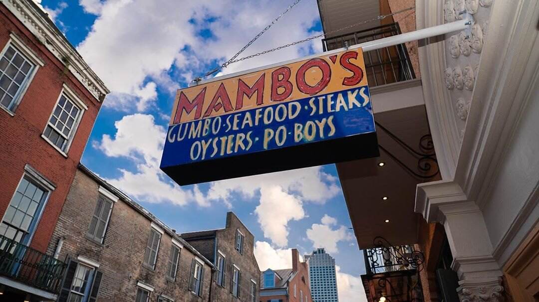 Mambo's hanging sign reading Gumbo, Seafood, Steaks, Oysters, Po-Boys against a blue Bourbon Street sky